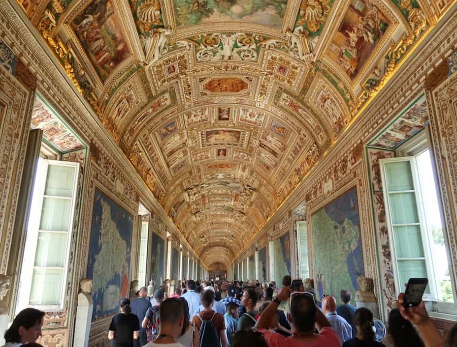 Hallway at the Vatican Museums showing crowds walking through with map tapestries either side and a elaborately decorated ceiling