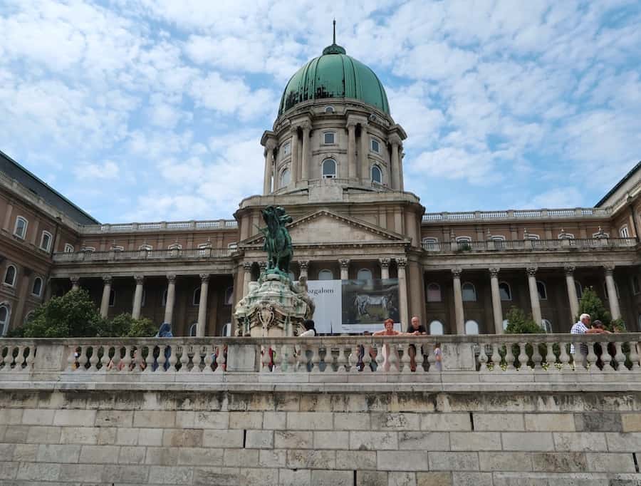 Image of Buda Castle in Budapest. The dome of the castle is green and there are people wondering around the courtyard below.