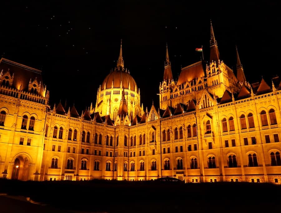 Image of the Hungarian Parliament Building at night. The building is very well lit and there are bird flying above.