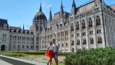 Feature Image for Life Of Y post - Best Things To Do On A Long Weekend In Budapest. Show Jeff and Zuzi standing on a low fall with the Hungarian Parliament Building behind them
