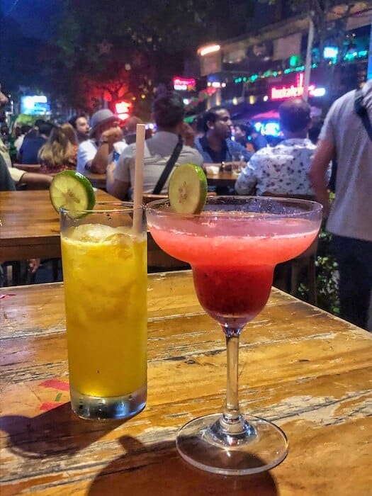 Image of a yellow and a pink cocktail with people in the background