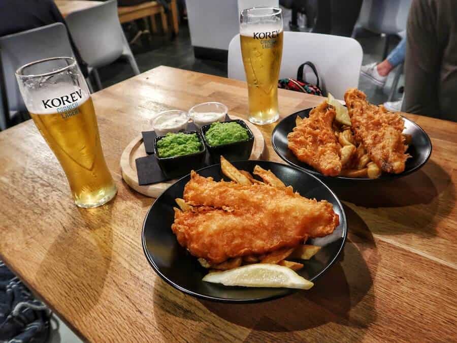 Two plates of battered fish and chips on a table with a couple of pints of lager