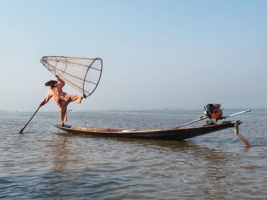 A fisherman at Inle Lake is balancing on the edge of his boat while holding a big wooden net