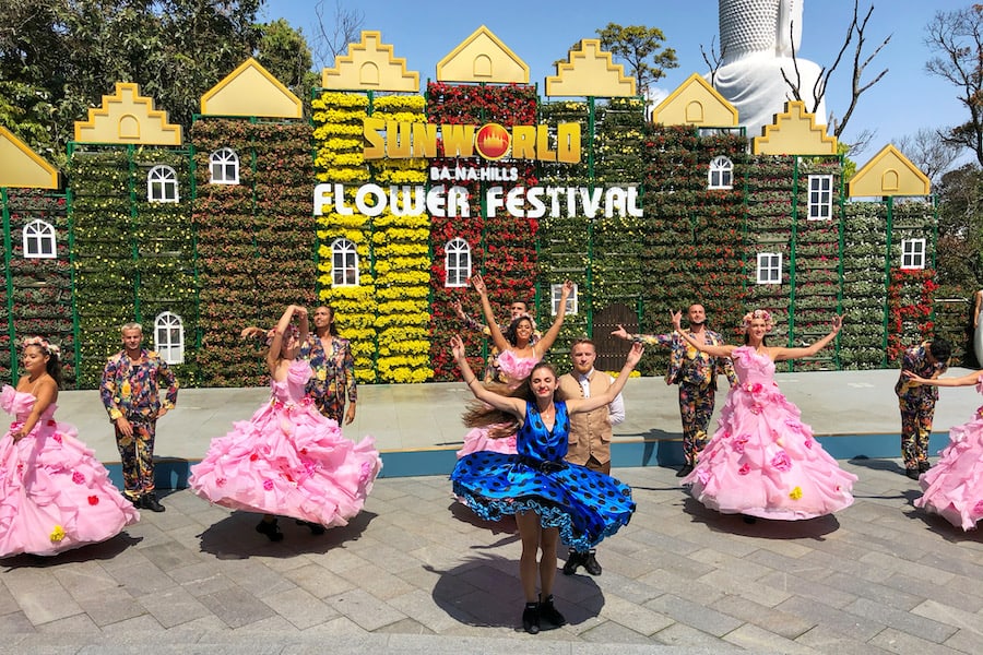 Image of Sunworld Ba Na Hills - Male and Female dancers in costume are dancing in front of a flower wall 