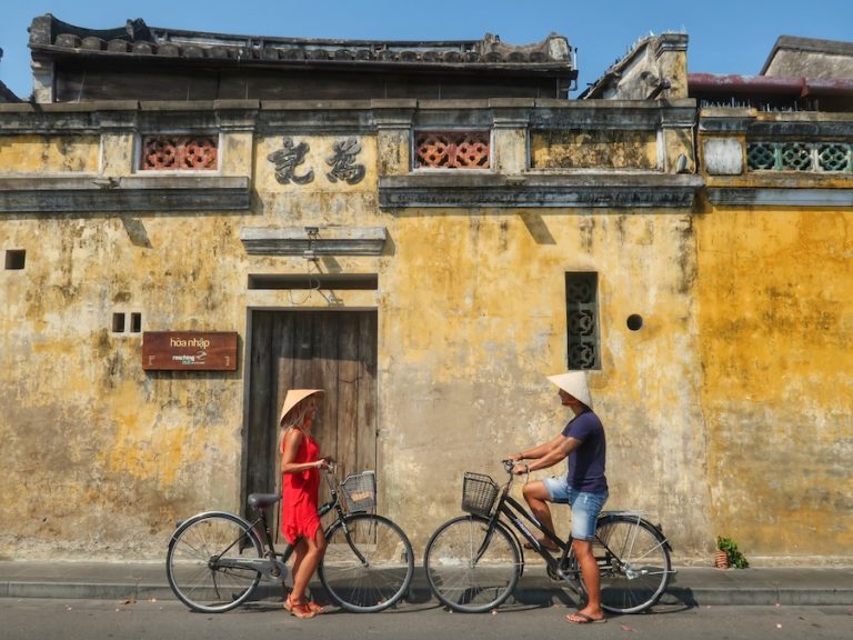 Title image for Is Vietnam Worth Visiting? Couple on bikes in Vietnam in front of an old yellow building.