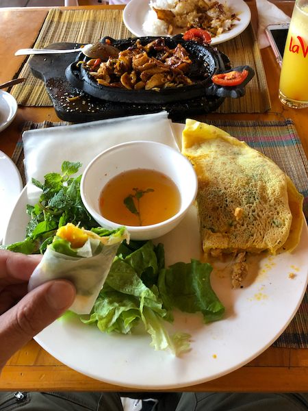 Image of some food from Vietnam. A spring roll is half eaten and there is a pancake wrap and some green on the plate with some meat in the middle