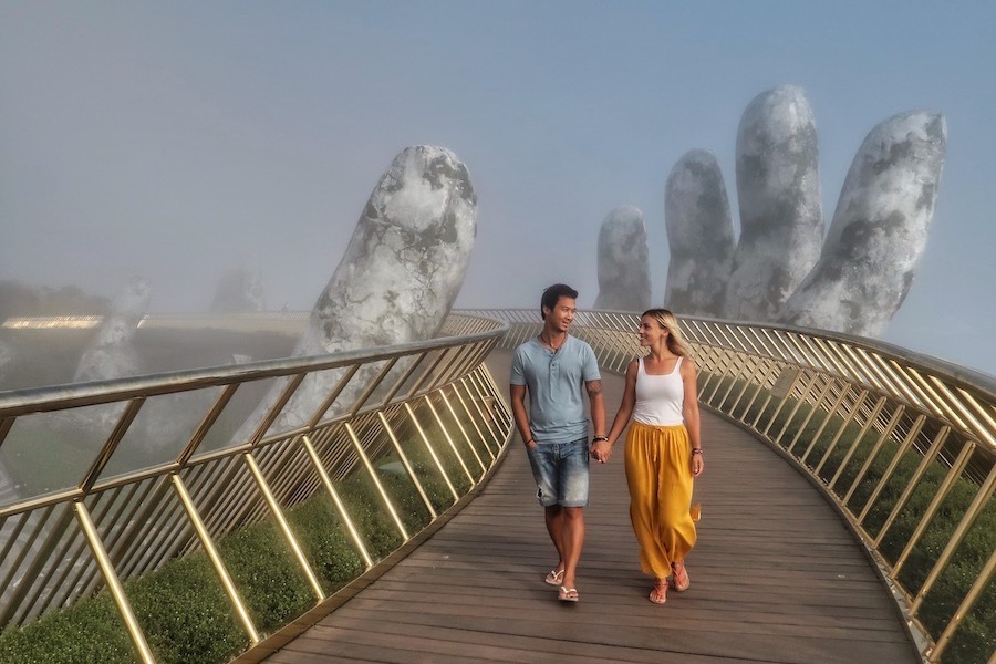 A couple walk on the bridge with a gold barrier and a giant stone hand in the background