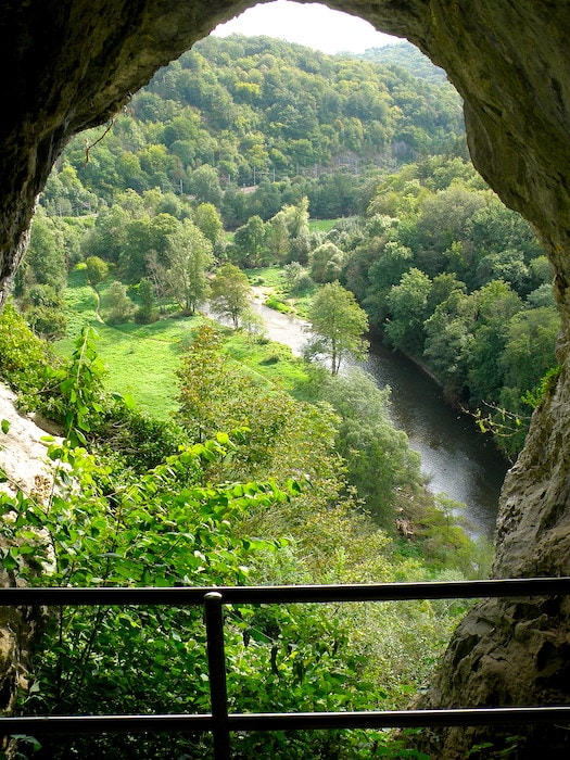 Image of Parc de Furfooz, the view is of a green forest and river from an archway in a cave