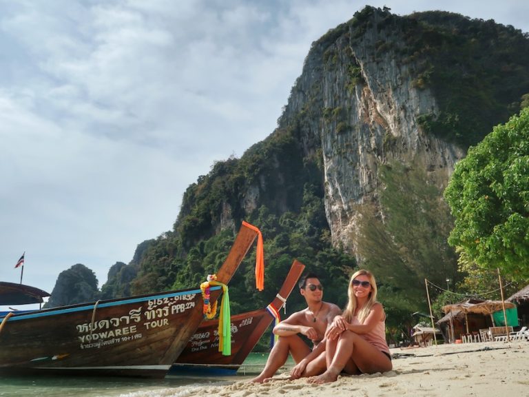 Cover image for Is Thailand Worth Visiting article showing couple sitting on a beach with long boats and mountains in the background