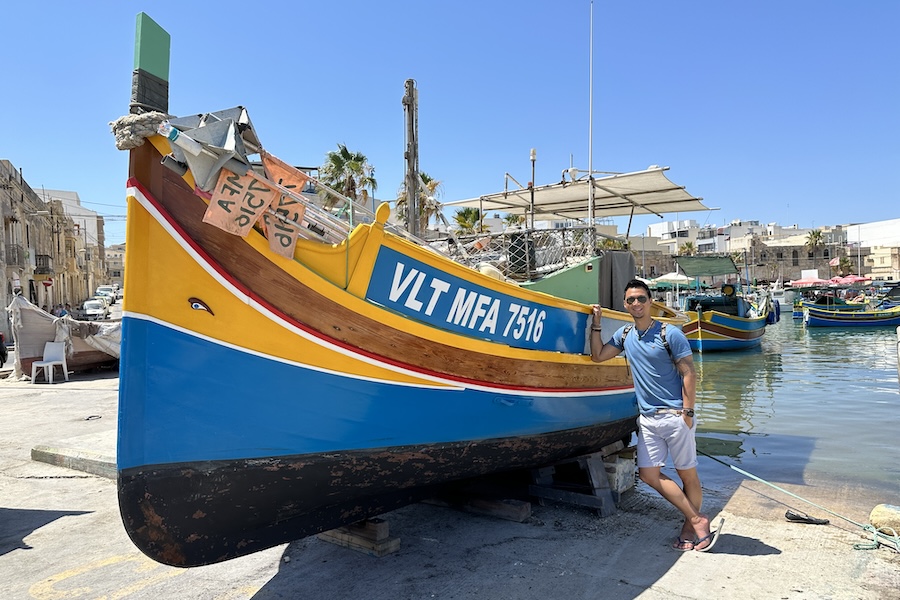 Man standing next to a traditional Maltese fishing boat in Marsaxlokk, Malta, on a sunny day"