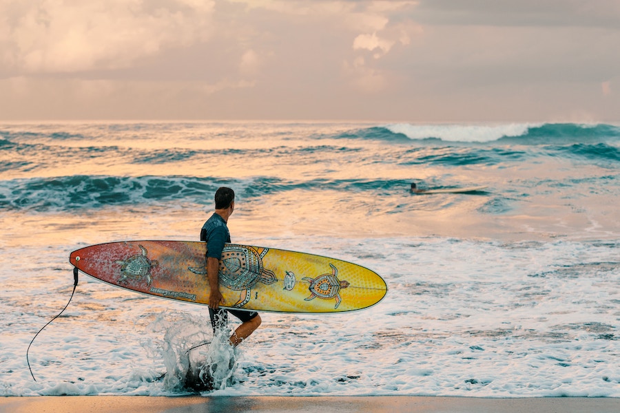 Man carrying a surfboard with a turtle pattern on it is walking in the water on a beach in Bali.