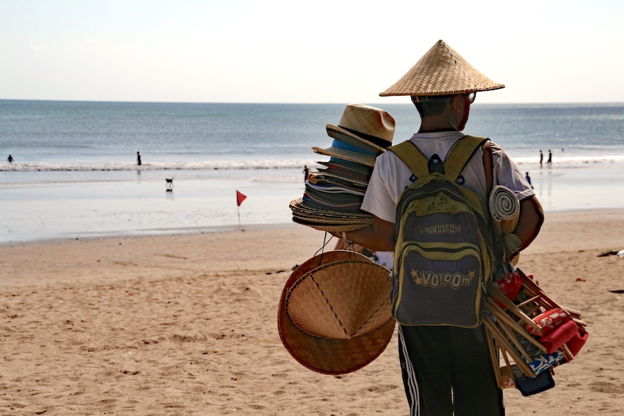 A hat vendor is on a beach in Bali carrying many types of hats.