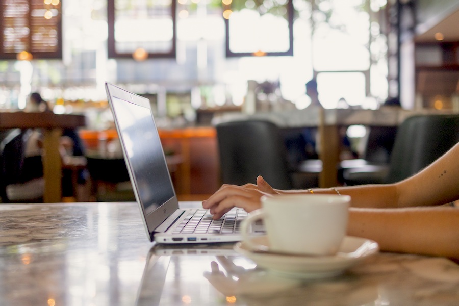 A laptop is open and woman is typing in a cafe with a coffee cup in the foreground.