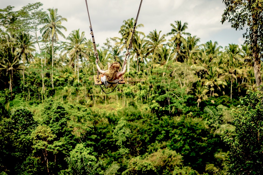 Woman in white dress is on a swing over lush vegetation and trees in Bali.