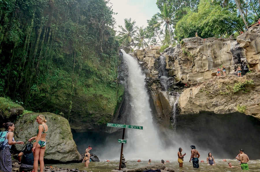 Tourists in the foreground at a tall waterfall in Bali surround by green vegetation.