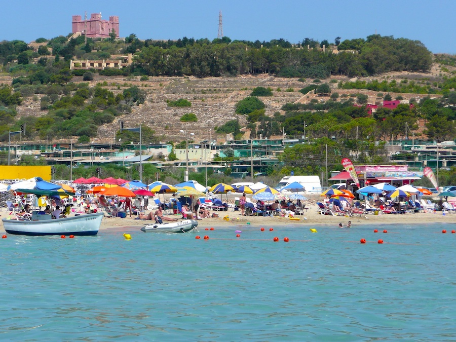 The beach front of Mellieha Bay in Malta with the sea in front and a red castle in the background