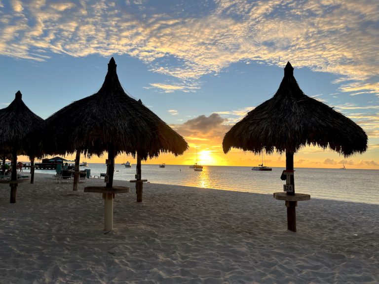 Cover image for Aruba sunset cruise showing a sunset at a beach in Aruba with sun umbrellas and boats in the sea in the background.