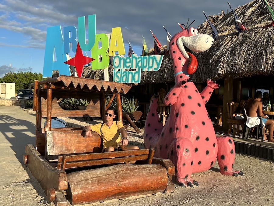 Sitting in a wooden cart next to a model dinosaur with colourful Aruba sign above me at Big Mamma Grill in Aruba.