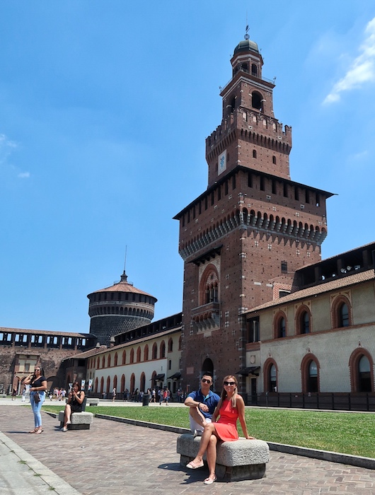 Jeff and Zuzi sit on a stone bench in a garden of the Castello Forzesco, an old castle. One of the towers is positioned behind the couple.