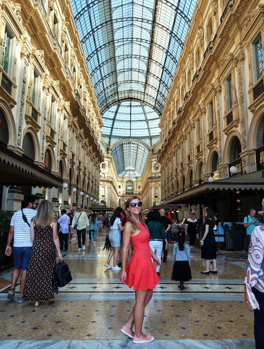 Zuzi in a red dress poses in the middle of a shopping centre in Milan. The Galleria is luxuriously decorated and has a high glass ceiling.