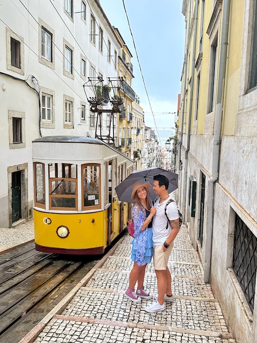 Jeff and Zuzi stand stand to a yellow tram under an umbrella in Lisbon Portugal